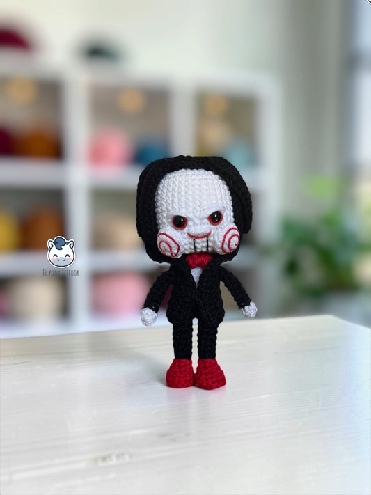 Amigurumi de Billy the Puppet inspirado en Saw, muñeco de crochet de terror para Halloween con traje negro, pajarita roja, mejillas en espiral y pelo negro.
