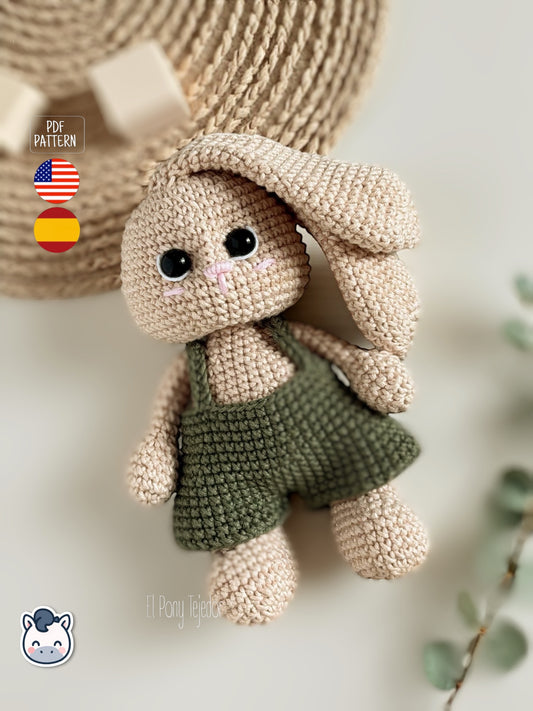 Patrón de ganchillo Amigurumi de un conejo color crema con peto verde oliva y orejas largas sostenido por una mano.Crochet Amigurumi pattern of a cream-colored bunny wearing olive green overalls and long ears held by a hand.