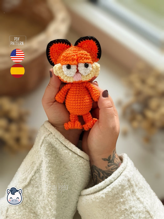 Amigurumi de gato naranja inspirado en Garfield tejido a ganchillo, con mejillas redondeadas, ojos bordados y rayas negras en la espalda.
Crochet orange cat amigurumi inspired by Garfield, featuring rounded cheeks, embroidered details and black stripes on the back.