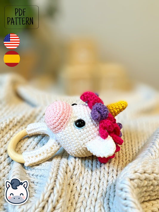 Patrón de ganchillo amigurumi de sonajero unicornio con cuerno dorado, melena de colores rosa y violeta sobre aro de madera. Crochet amigurumi pattern of a baby unicorn rattle with a golden horn, pink and purple mane on a wooden ring.