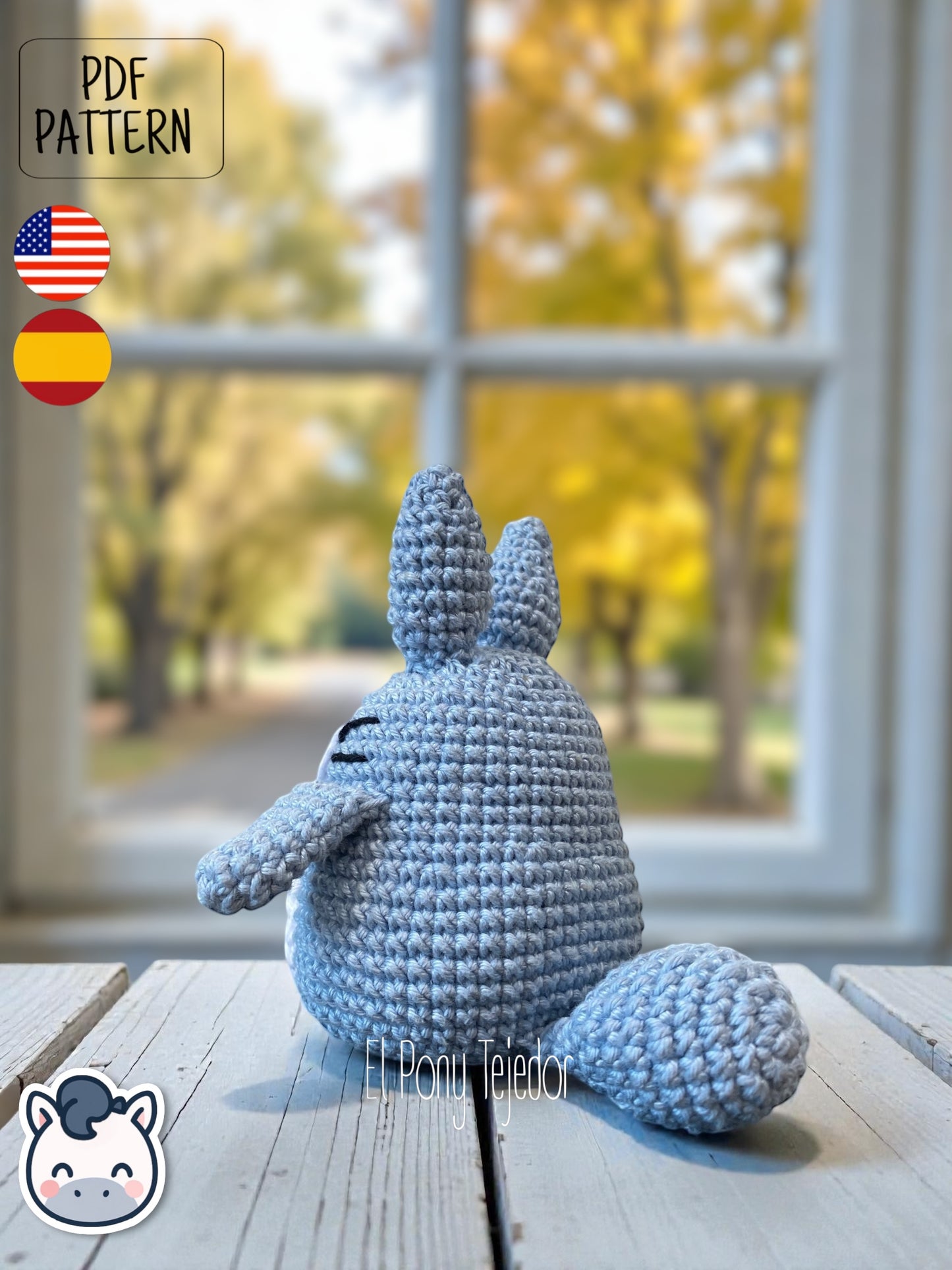 Amigurumi de Totoro de ganchillo en color azul y blanco sobre una mesa de madera con fondo de bosque otoñal. Blue and white crochet Totoro amigurumi on a wooden table with a blurry autumn forest background.
