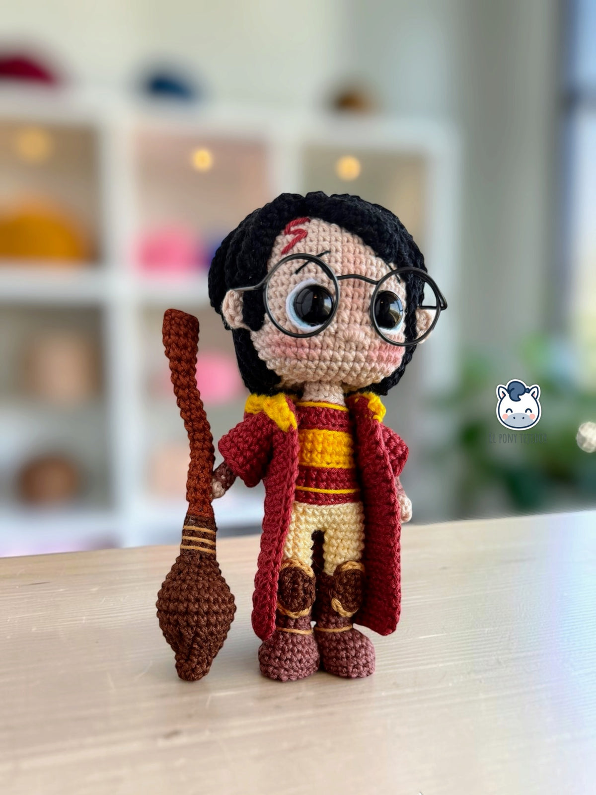 Amigurumi de Harry Potter con uniforme rojo y amarillo de Quidditch, sosteniendo la escoba Nimbus 2000 de crochet, visto en tres cuartos.