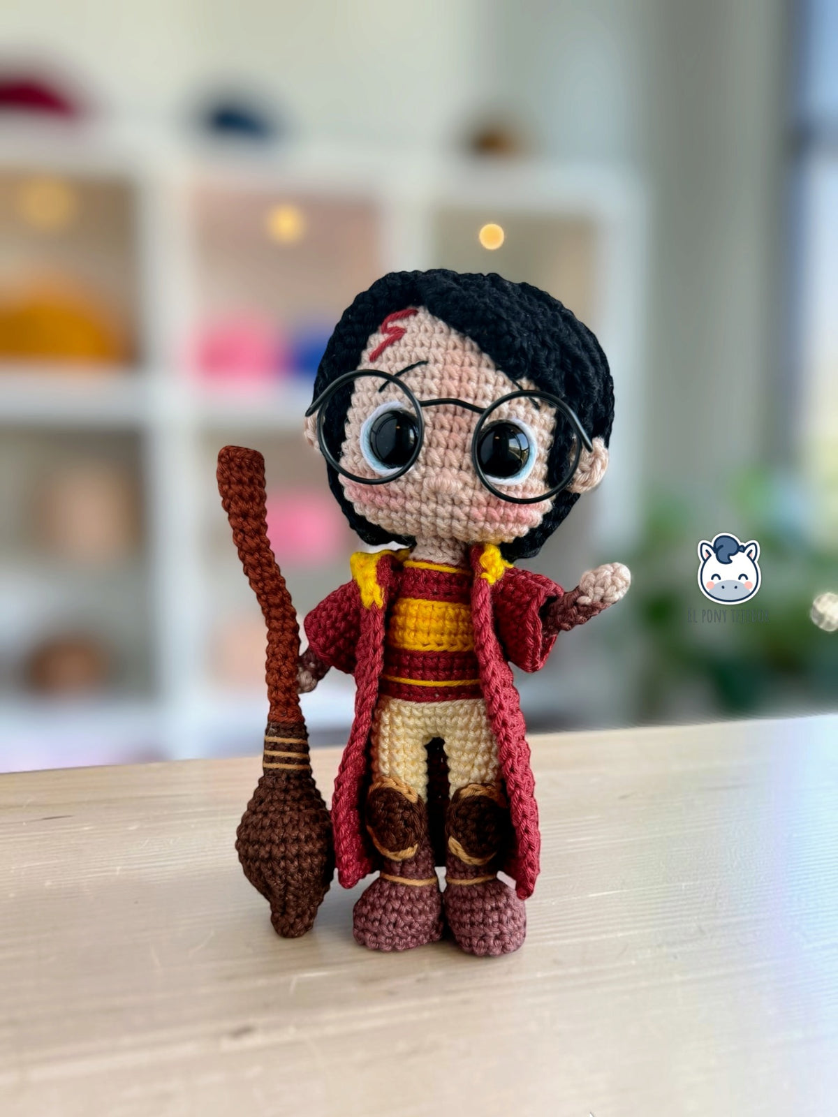 Amigurumi de Harry Potter con gafas y uniforme de Quidditch, visto de frente con la escoba Nimbus 2000 de crochet y una mano levantada.