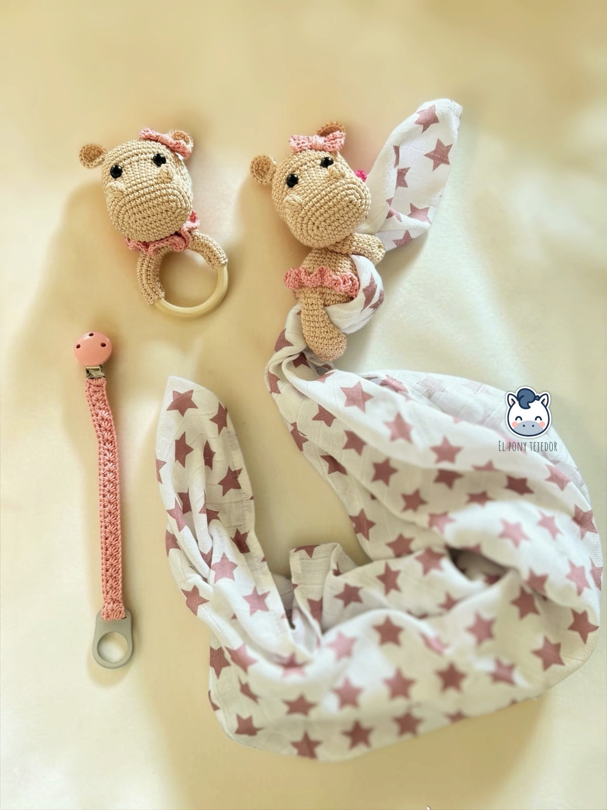 Pack de hipopótamos amigurumi beige y rosa, con aro, tela blanca de estrellas rosas y cinta de crochet con pinza, sobre una manta clara.