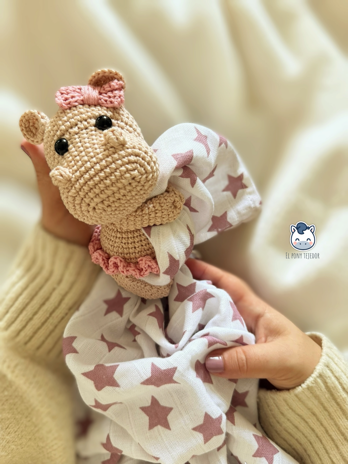 Pack de hipopótamos amigurumi beige y rosa, con aro, tela blanca de estrellas rosas y cinta de crochet con pinza, sobre una manta clara.