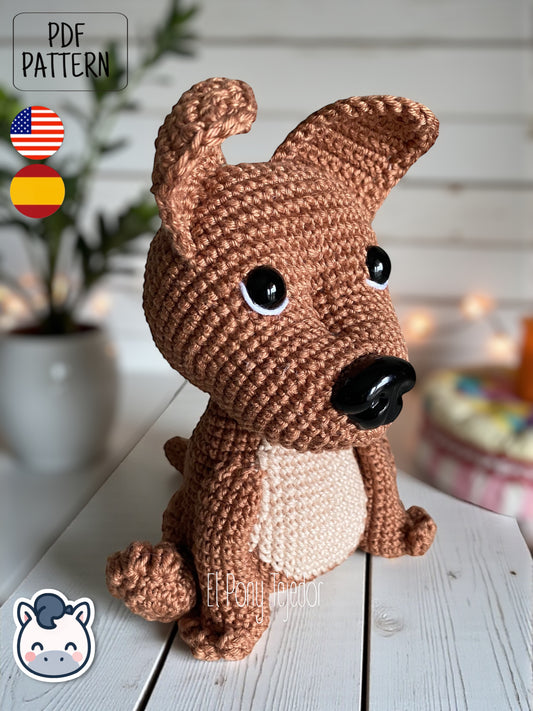 Patrón de ganchillo Amigurumi de perro American Stafford (Pitbull) de color marrón y beige sentado sobre una superficie de madera. Crochet Amigurumi pattern of an American Staffordshire dog (Pitbull) in brown and beige sitting on a white wooden surface.