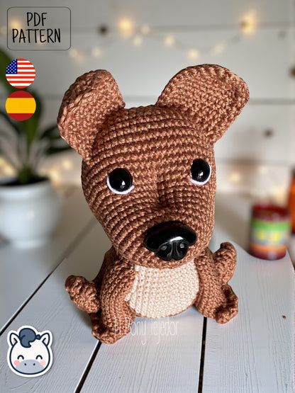 Patrón de ganchillo Amigurumi de perro American Stafford (Pitbull) de color marrón y beige sentado sobre una superficie de madera. Crochet Amigurumi pattern of an American Staffordshire dog (Pitbull) in brown and beige sitting on a white wooden surface.