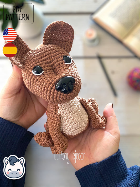 Patrón de ganchillo Amigurumi de perro American Stafford (Pitbull) de color marrón y beige sentado sobre una superficie de madera. Crochet Amigurumi pattern of an American Staffordshire dog (Pitbull) in brown and beige sitting on a white wooden surface.