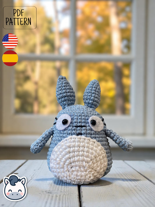 Amigurumi de Totoro de ganchillo en color azul y blanco sobre una mesa de madera con fondo de bosque otoñal. Blue and white crochet Totoro amigurumi on a wooden table with a blurry autumn forest background.