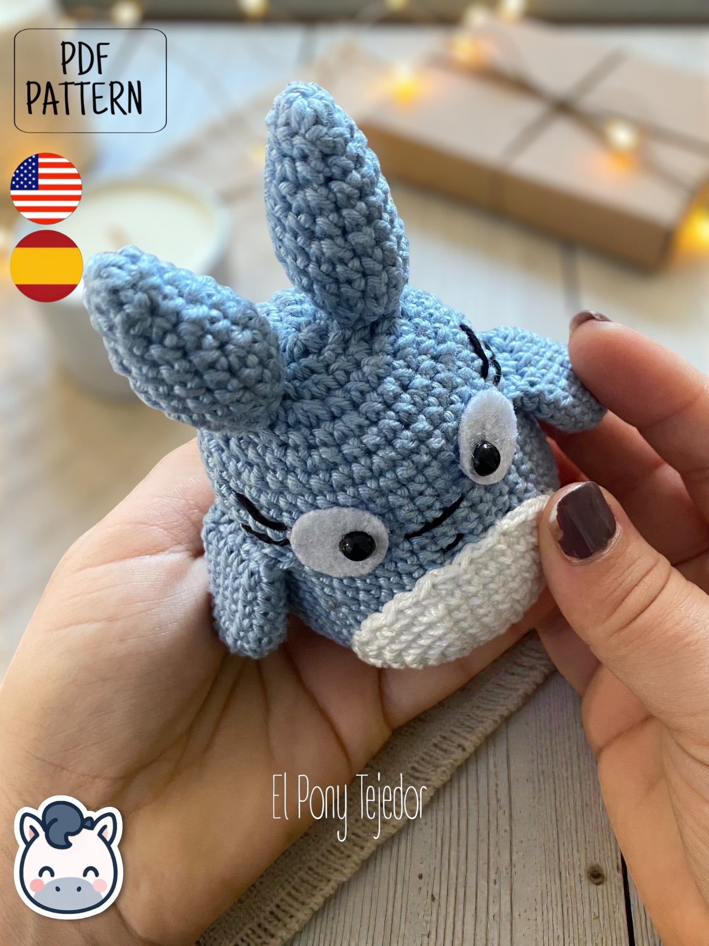 Amigurumi de Totoro de ganchillo en color azul y blanco sobre una mesa de madera con fondo de bosque otoñal. Blue and white crochet Totoro amigurumi on a wooden table with a blurry autumn forest background.