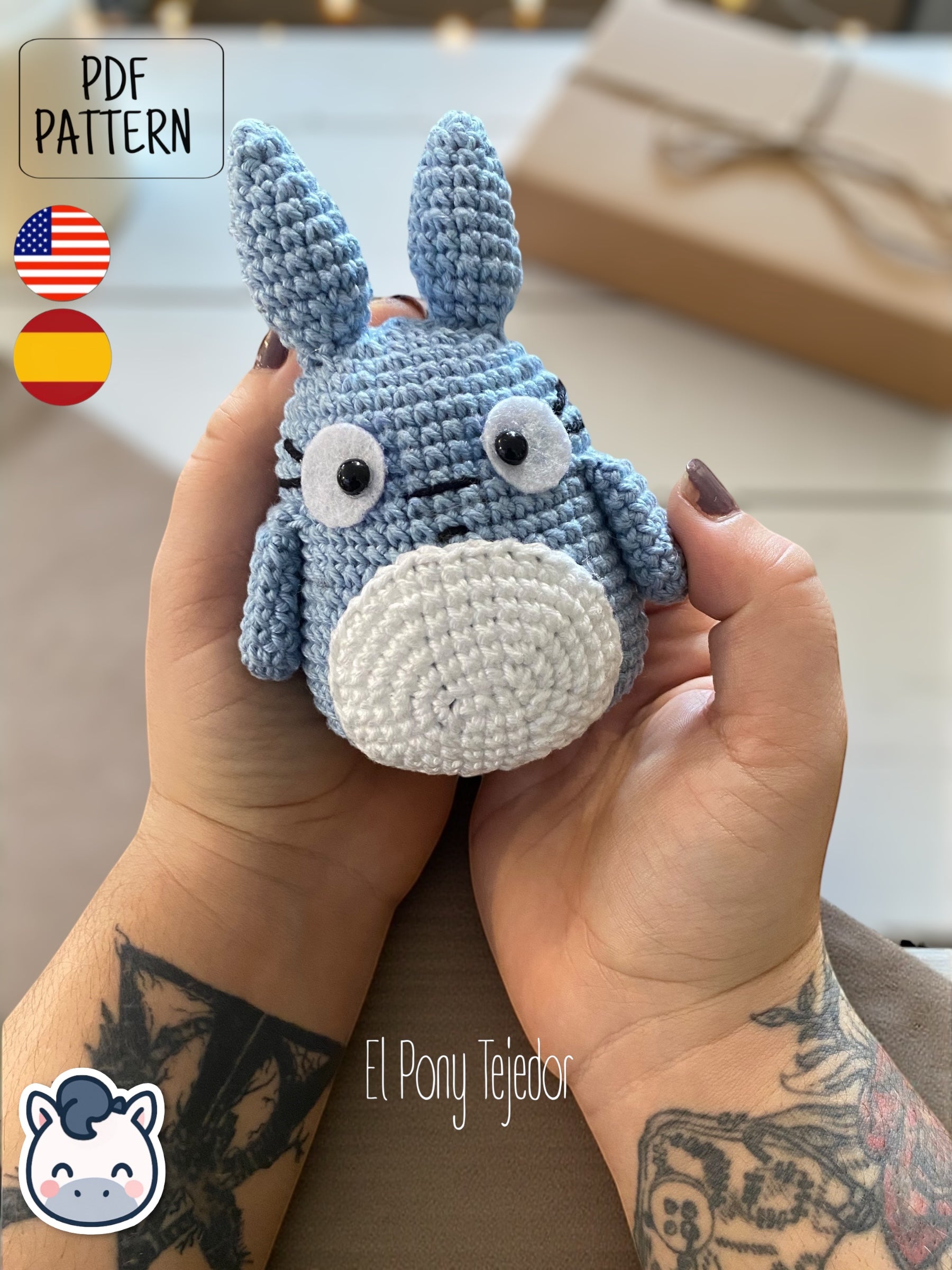 Amigurumi de Totoro de ganchillo en color azul y blanco sobre una mesa de madera con fondo de bosque otoñal. Blue and white crochet Totoro amigurumi on a wooden table with a blurry autumn forest background.