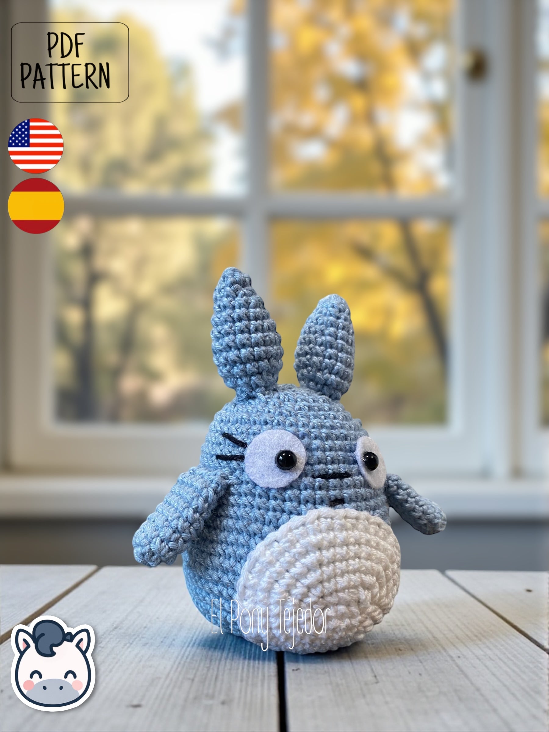 Amigurumi de Totoro de ganchillo en color azul y blanco sobre una mesa de madera con fondo de bosque otoñal. Blue and white crochet Totoro amigurumi on a wooden table with a blurry autumn forest background.