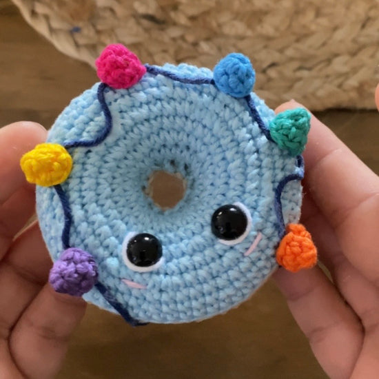 Video tutorial y Patrón de ganchillo amigurumi de dos donuts navideños: uno beige con glaseado blanco y acebo, y otro azul con una guirnalda de luces de colores.  Video tutorial Crochet amigurumi pattern of two Christmas donuts: one beige with white icing and holly, and one blue with a colorful string of Christmas lights